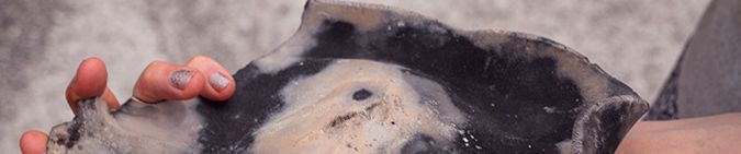 Hände und Unterarme einer Person, in der Linken Hand (Bildvordergrund) hält sie ein gebranntes, flaches Keramikobjekt dass sich passgenau an die Handfläche anschmiegt und über dem Ballen und am Handgelenk aufliegt. Im vorderen Teil der Hand ist es gleich