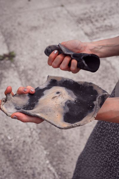 Hände und Unterarme einer Person, in der Linken Hand (Bildvordergrund) hält sie ein gebranntes, flaches Keramikobjekt dass sich passgenau an die Handfläche anschmiegt und über dem Ballen und am Handgelenk aufliegt. Im vorderen Teil der Hand ist es gleich