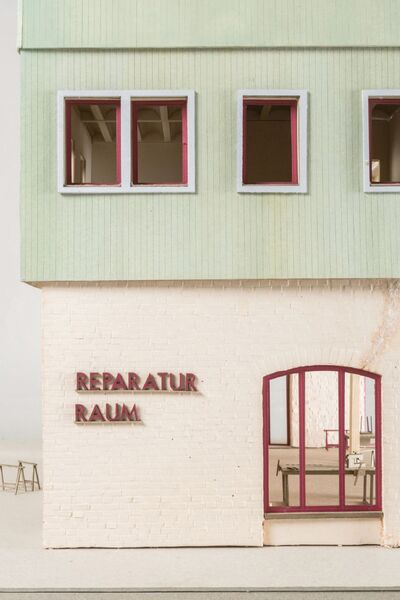 Ein Foto eines farbigen Modells mit beigem/weißen geziegelten Erdgeschoß, Salbei farbenen hölzernen 1. Obergeschoß. Roter Schriftzug auf dem "Reparatur Raum" steht. Rote Fensterrahmen.