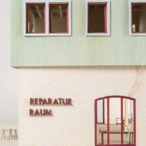 Ein Foto eines farbigen Modells mit beigem/weißen geziegelten Erdgeschoß, Salbei farbenen hölzernen 1. Obergeschoß. Roter Schriftzug auf dem "Reparatur Raum" steht. Rote Fensterrahmen.